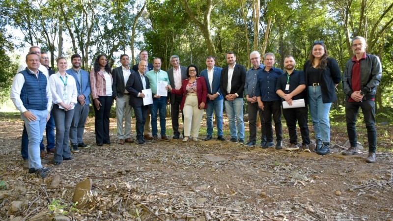Imagem de um grupo de pessoas de pé, olhando para a câmera. Eles estão em um terreno com grama seca no chão e árvores ao fundo.