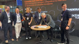 Grupo de pessoas em torno de uma pequena mesa onde é assinado um documento.