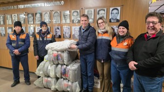 Um grupo de pessoas trajando uniformes da Defesa Civil do RS e outros dois homens vestindo jaqueta de naylon posam para a foto ao lado de uma pilha embalagens de cobertores