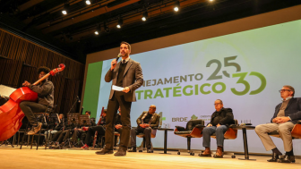 Um home fala ao microfone num palco com pessoas sentadas em uma fileira atrás