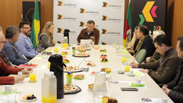 Foto de um grupo de pessoas sentados ao redor de uma longa mesa de reuni&otilde;es. H&aacute; itens de caf&eacute; da manh&atilde; na mesa