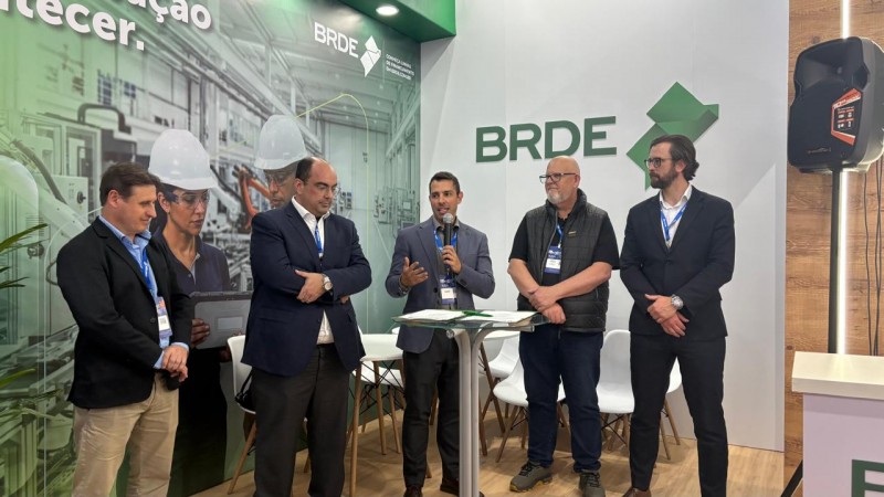 Cinco homens brancos est&atilde;o em p&eacute; no estande do BRDE durante um evento. Ao centro, um deles fala ao microfone ao lado de uma mesa alta com documentos. Ao fundo, h&aacute; o logotipo do BRDE e um painel com imagens de ambiente industrial e trabalhadores utilizando equipamentos de prote&ccedil;&atilde;o. O grupo veste trajes empresariais e crach&aacute;s de identifica&ccedil;&atilde;o. O espa&ccedil;o conta com cadeiras brancas ao fundo, ilumina&ccedil;&atilde;o interna e estrutura de feira de neg&oacute;cios.