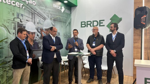 Cinco homens brancos est&atilde;o em p&eacute; no estande do BRDE durante um evento. Ao centro, um deles fala ao microfone ao lado de uma mesa alta com documentos. Ao fundo, h&aacute; o logotipo do BRDE e um painel com imagens de ambiente industrial e trabalhadores utilizando equipamentos de prote&ccedil;&atilde;o. O grupo veste trajes empresariais e crach&aacute;s de identifica&ccedil;&atilde;o. O espa&ccedil;o conta com cadeiras brancas ao fundo, ilumina&ccedil;&atilde;o interna e estrutura de feira de neg&oacute;cios.
