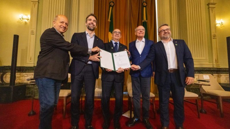 Foto de cinco homens de terno, lado a lado, posando para a foto. Eles seguram um documento aberto.