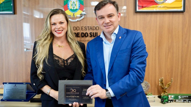 Foto mostra um homem e uma mulher sorrindo posando para a foto segurando uma documento/convite