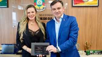 Foto mostra um homem e uma mulher sorrindo posando para a foto segurando uma documento/convite